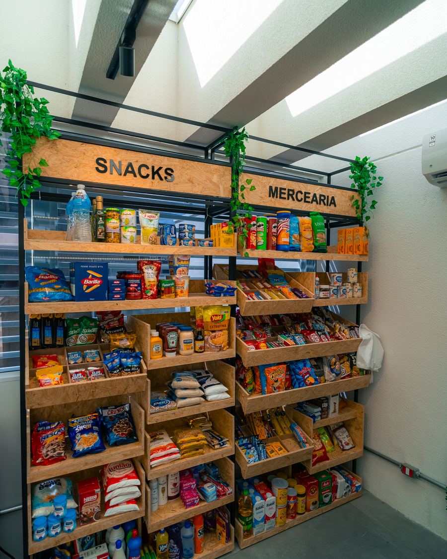 Loja autônoma completa ocupando o canto de uma sala pequena, com duas gôndolas de madeira cheias de mercearia, snacks, higiene e limpeza.