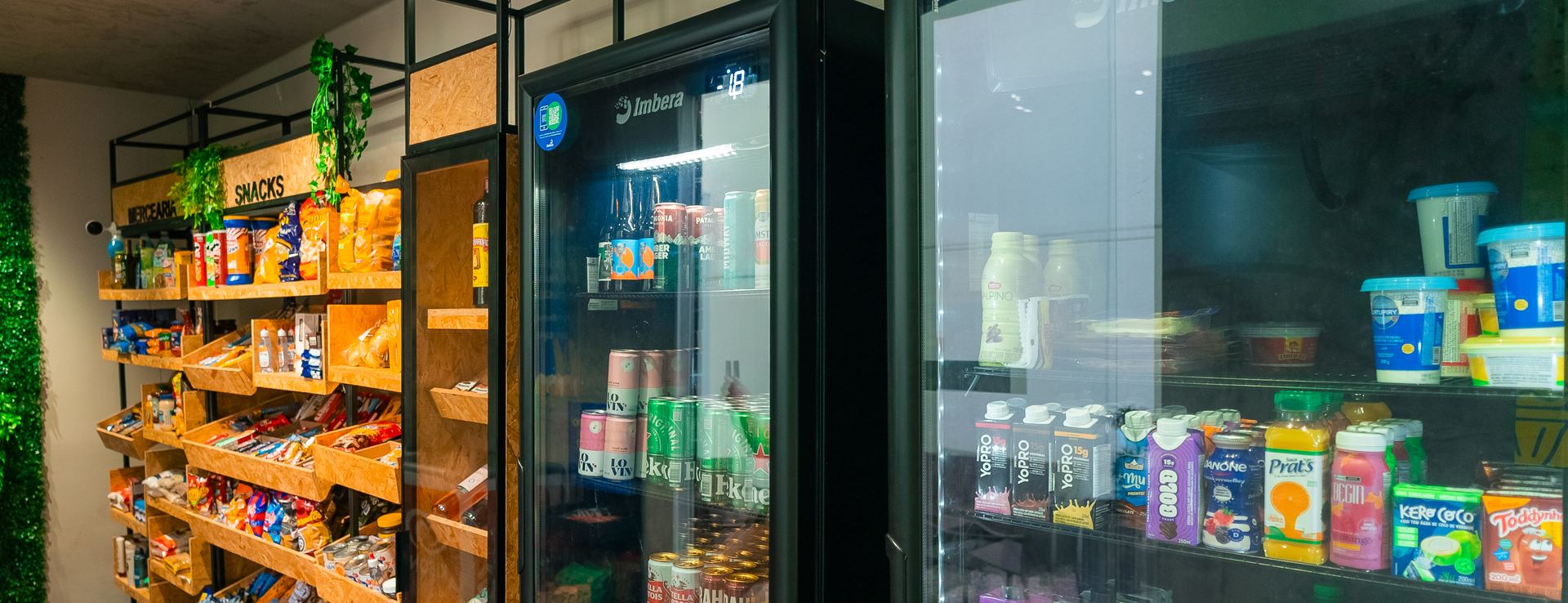 Interior de uma loja autônoma montada: prateleiras de mercearia e snacks à esquerda e duas geladeiras de porta de vidro com bebidas e laticínios à direita.
