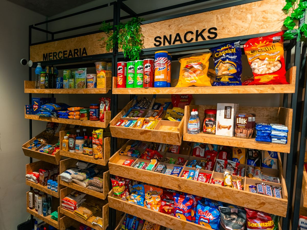 Gôndola de madeira vista de frente, cheia, com as seções de mercearia e snacks sinalizadas e uma câmera de segurança no alto à esquerda.